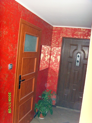 Czerwone ściany z dekoracyjną fakturą w przedpokoju, drewniane drzwi z szybą, ciemne drzwi wejściowe z ozdobnym szkłem, obraz na ścianie i roślina doniczkowa na podłodze.